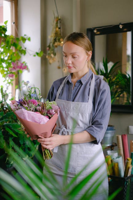 A young woman with fair skin and light brown hair tied back, wearing a grey shirt and light grey apron, is holding a bouquet of fresh flowers in a floral shop. The bouquet includes pink roses, purple 