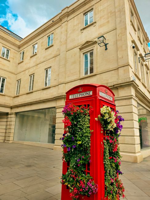 A vintage red telephone booth situated on a paved city street corner, adorned with an abundant display of colorful flowers and lush greenery cascading from the top and sides. The flowers include pink,