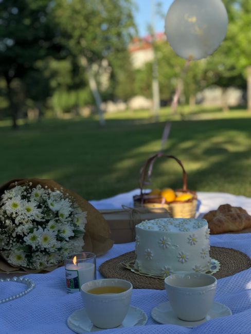 A floral arrangement featuring a bouquet of small white daisy-like flowers with yellow centers, wrapped in brown paper, resting on a white tablecloth outdoors. The scene includes a small, lit candle i