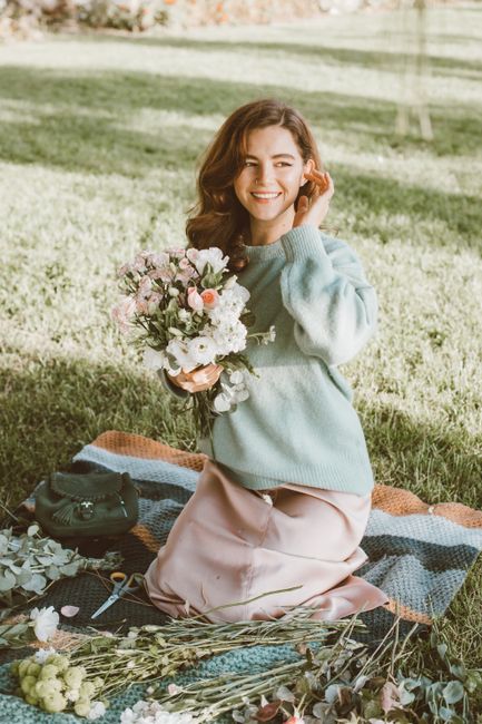 A young woman with wavy brown hair smiles warmly while kneeling on a blanket in a park, holding a vibrant bouquet of fresh flowers including pink roses, white peonies, and greenery. She is wearing a l