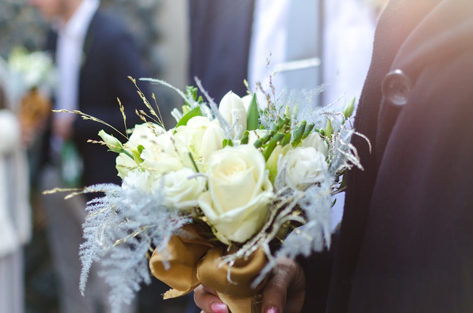 A close-up view of a wedding bouquet featuring cream-white roses with lush, velvety petals, accented by delicate fern-like foliage and wispy, frosted-looking decorative elements. The bouquet is wrappe