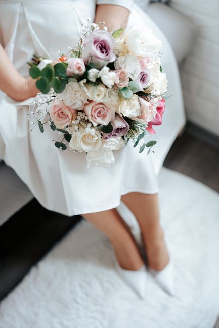 A bride sitting on a white cushion in a softly lit room, holding a large, elegant bouquet arrangement of fresh flowers. The bouquet features predominantly pastel pink and white roses, complemented by 