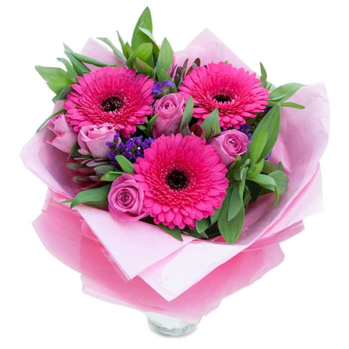 I'd describe this bouquet as a balance of playful colour and considered form, conveyed here in words so someone who can't see it can still sense its mood. Three vibrant Gerbera daisies in saturated fuchsia take centre stage, their petals splayed like tiny suns around deep, dark centres that add a sophisticated contrast. Nestled nearby are roses in a softened lavender-pink or dusty-rose tone, their tightly furled petals offering the feel of newness and intimacy rather than full flourish. Dainty clusters of purple statice or limonium peek through the spaces between the larger blooms, lending a textured, wildflower note that keeps the arrangement from feeling too formal. The greenery is abundant and tactile-broad salal-like leaves and slender eucalyptus stems that bring a cool, slightly citrusy-green aroma and a crisp visual frame. The wrapping is worth noting: an inner mattress of pale pink tissue paper cushions the stems, while an overwrap of translucent, slightly crinkled fuchsia-pink cellophane gives a soft shimmer and unifies the pink palette; thoughtful folds hold the bouquet like a well-made present. The clear glass vase rim is visible at the bottom, signalling freshness and immediate display potential. In the kind of soft daylight common to homes around Colliers Wood and nearby Merton, the bouquet reads as cheerful and affectionate-perfect for celebrating milestones, sending thanks, or surprising someone just because-crafted with the steady hand of a neighbourhood florist who values scent, texture, and honest colour.